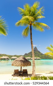 Two Sun Lounger Chairs Under A Thatched Parasol On A Sand Beach With A View On The Lagoon And Mount Otemanu On The Tropical Island Of Bora Bora, Near Tahiti, In French Polynesia.