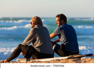 Two Surfers Sitting On Their Surf Boards On The Beach Discussing The Waves