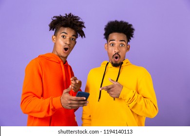 Two Surprised African Man Friends Dressed In Colorful Hoodies Standing Isolated Over Violet Background, Pointing At Mobile Phone