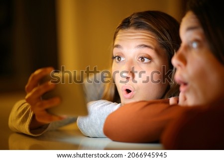 Two surprised friends in the night at home checking smart phone content