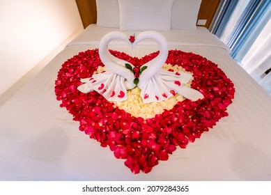 Two Swans Made From Towels Are Kissing On Honeymoon White Bed. Creamy Pillow And Heart Form, Valentine Signature Made From Red Rose Flower On Bed Decoration In Bedroom. Honey Moon