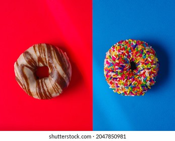 Two Sweet Donuts Covered With Icing, Chocolate And Pastry Sprinkles On A Red And Blue Background. Minimalism. There Are No People In The Photo. Sweet Food, Lots Of Calories, Birthday, Holiday.