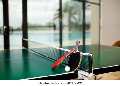 Two table tennis or ping pong rackets and balls on a green table with net, swimming pool, palm tree and sea in the background; shallow DOF, focus on rackets