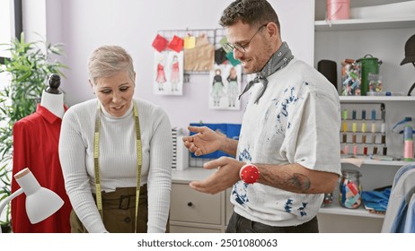 Two tailors, a woman and a man, interact in a well-organized tailor shop with garments and design sketches visible. - Powered by Shutterstock