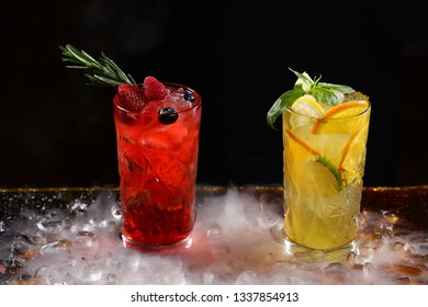 Two tall crystal glasses with lemonade made from orange, lime, strawberries, blueberries and raspberries on a black background and pieces of ice on the table. Nitrogen vapor in the frame.