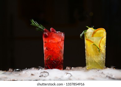 Two tall crystal glasses with lemonade made from orange, lime, strawberries, blueberries and raspberries on a black background and pieces of ice on the table. Nitrogen vapor in the frame. 