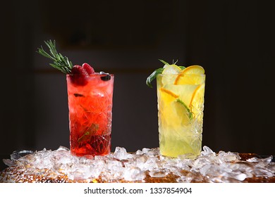 Two tall crystal glasses with orange, lime, strawberry, blueberry and raspberry lemonades on a black background and with pieces of ice on the table. Still life concept.