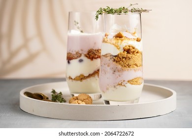 Two tall glasses with a layered creamy yoghurt dessert trifle with cookies, amaretti, cherries and thyme in a tropical setting