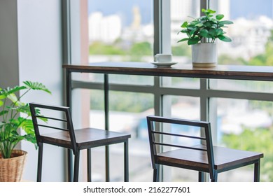 Two Tall Industrial Loft Design Steel Wooden Chairs Placed Near Table Counter With Tree Pot And White Hot Coffee Cup In Company Living Room In Front Glass Window Show Blurred City View In Background.