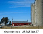 two tall silos next to a one story red barn on a farm in winter with a bright blue sky in the background