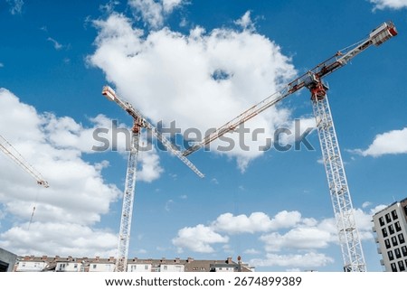 Two tall tower cranes at a construction site near contemporary apartment blocks. Concept of city growth, real estate investment, housing development, and modern infrastructure.