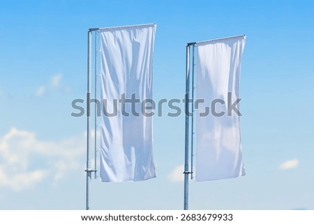 Two tall vertical white flags waving on metal poles against clear blue sky, minimalistic outdoor advertising concept