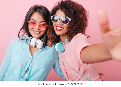 Two Tanned Ladies With Headphones Making Selfie During Indoor Party. Enthusiastic Curly Woman In Sunglasses Taking Picture Of Herself Near Romantic Asian Girl In Blur Cotton Shirt.