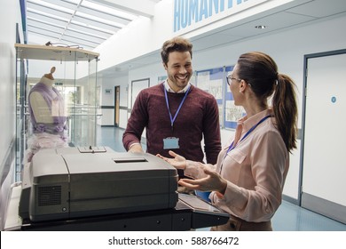 Two Teachers Are Talking While Standing At A Printer In The School Hall. 