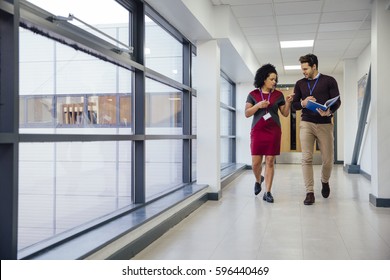 Two Teachers Are Walking And Talking Together Down The School Hall. They Are Discussing A Student's Work. 