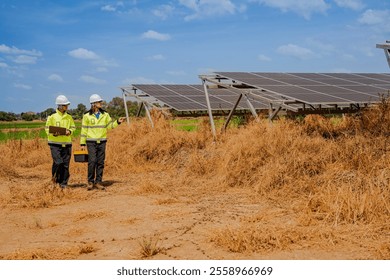 Two technicians in high-visibility jackets and helmets walk through a solar farm, carrying tools and a laptop, inspecting photovoltaic panels surrounded by dry grass under a clear sky.