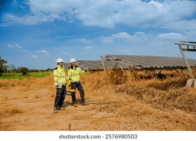 Two technicians in high-visibility jackets and helmets walk through a solar farm, carrying tools and a laptop, inspecting photovoltaic panels surrounded by dry grass under a clear sky.
