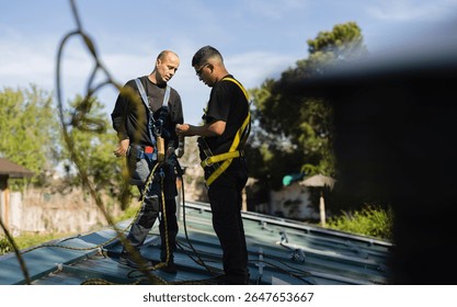 Two technicians are preparing their safety harnesses and ropes before working on photovoltaic panels installed on a metal roof, ensuring workplace safety during a sunny day