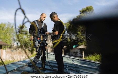 Two technicians are preparing their safety harnesses and ropes before working on photovoltaic panels installed on a metal roof, ensuring workplace safety during a sunny day