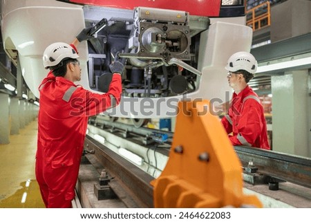 Two technicians in red overalls communicate via walkie-talkies while working on the coupling system of a train in a maintenance facility.