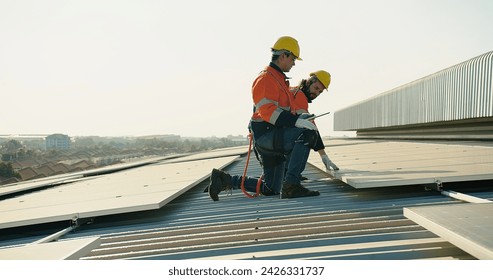 Two technicians in safety harnesses are working on the installation of solar panels on a sunny rooftop against a clear sky background