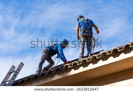 Two technicians working on rooftop wearing safety equipment are installing photovoltaic panels on a residential building
