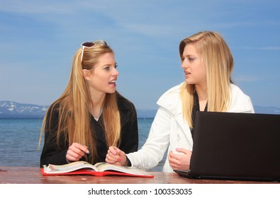 Two teen aged girl students sitting at a park bench studying with a book and computer.