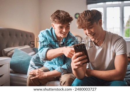 Two Teenage Boys At Home In Bedroom Looking At Social Media On Mobile Phone And Laughing