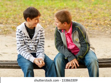 Two Teenage Friends Talking While Sitting On The Bench In The Park