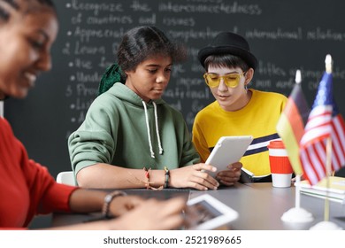 Two teenage girls collaborating on project and using digital tablet while sitting at table during lesson in school classroom - Powered by Shutterstock