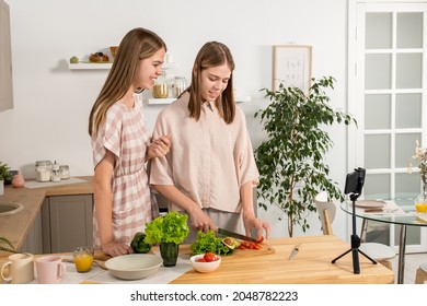 Two Teenage Girls Cooking Vegetable Salad By Kitchen Table During Livestream