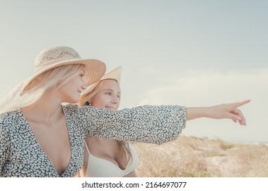 Two Teenage Girls Enjoy A Sunny Day On The Mediterranean Coast During Their Summer Vacation. They Look Far Away Pointing Something At The Horizon. Friendship Concept.