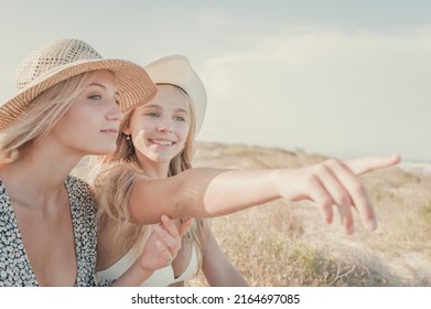 Two Teenage Girls Enjoy A Sunny Day On The Mediterranean Coast During Their Summer Vacation. They Look Far Away Pointing Something At The Horizon. Friendship Concept.