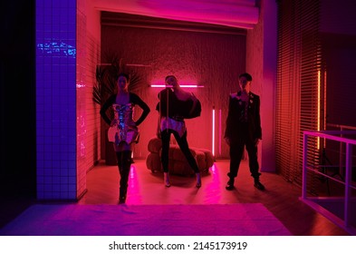 Two teenage girls and guy in posh stage costumes making performance during vogue ball in spacious room or studio lit by neon light