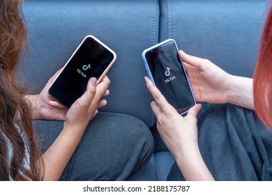 Two Teenage Girls With Phones In Their Hands Are Sitting At Home On Bed. Social Media Tiktok Is Loaded On Screen.