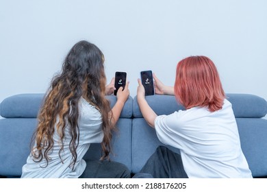 Two Teenage Girls With Phones In Their Hands Are Sitting At Home On Bed. Social Media Tiktok Is Loaded On Screen.