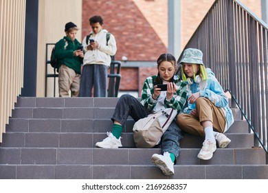 Two teenage girls sitting on stairs outdoors and watching video on mobile phone with young boys in background - Powered by Shutterstock