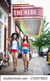 Two Teenage Girls Walking Down A Small Town Main Street By A Theater 