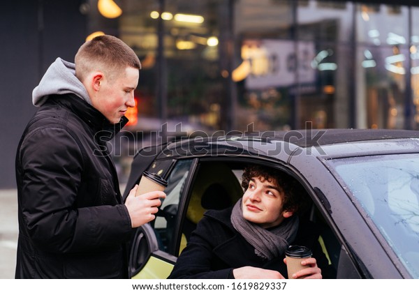 Two teenage youth students boy drink coffee. One\
of them standing near car drinking hot drink. Friends say during\
the meeting and hold paper cups, sits in car with coffee near\
business center.