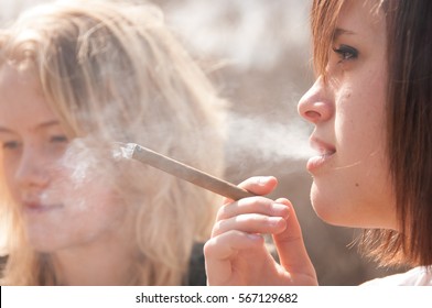 Two Teenagers Smoking Thin Cigars Outside In The Sun.  Cigar Smoke Backlit, Coming Out Of Her Mouth And Hands In The Air.  Blonde And Brunette Girls Smoking Cigars Outside In The Sun.