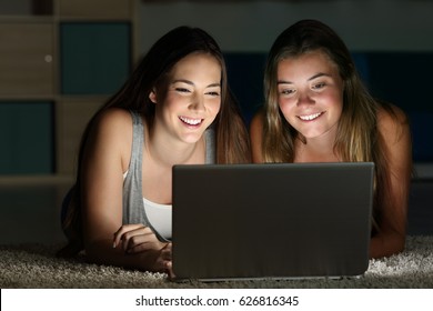 Two teens watching on line content in a laptop lying on the floor in a room at home with a dark light in the background