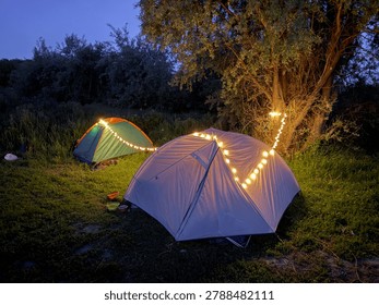 Two tents lit with string lights at dusk. Grass covers ground beneath tents. Trees stand tall behind camping setup. No animals or plants clearly visible