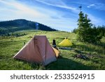 Two tents, orange and yellow, on grassy hillside under clear blue sky. Camping gear scattered nearby, distant smoke plume rises from fire. Surrounded by rolling hills and dense pine trees.