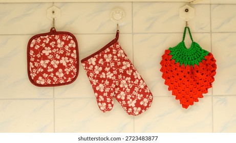 two textile potholders with a floral pattern and one knitted potholder in the shape of a strawberry hang on white hooks against a light tiled background in the kitchen interior