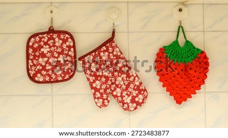two textile potholders with a floral pattern and one knitted potholder in the shape of a strawberry hang on white hooks against a light tiled background in the kitchen interior