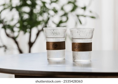 Two textured clear glasses with wooden bands sit on a table, greenery blurred in the background. - Powered by Shutterstock