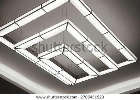 Two tiered, rectangular modern chandeliers creating abstract, nested geometric patterns. Contemporary interior ceiling light design with clean, bright white glow.
