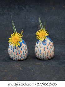 Two tiny hand-painted ceramic bud vases with colorful dash patterns holding bright yellow dandelions. Miniature egg-shaped artisanal pottery on a dark textured slate background. Concept of summer mood, playful home decor, and creative handmade gifts. High-quality still life photography for lifestyle blogs and interior design.
