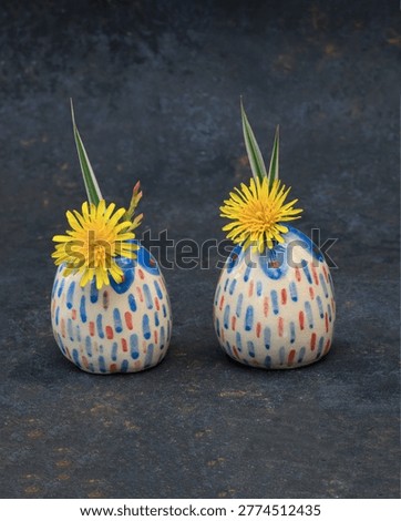 Two tiny hand-painted ceramic bud vases with colorful dash patterns holding bright yellow dandelions. Miniature egg-shaped artisanal pottery on a dark textured slate background. Concept of summer mood, playful home decor, and creative handmade gifts. High-quality still life photography for lifestyle blogs and interior design.
