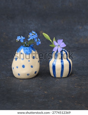 Two tiny hand-pinched ceramic bud vases with blue polka dots and vertical stripes, holding small wildflowers. Miniature artisanal pottery with organic irregular shapes on a dark textured background. Concept of slow-made craft, unique handmade gifts, and delicate floral aesthetics for modern interior decor.
